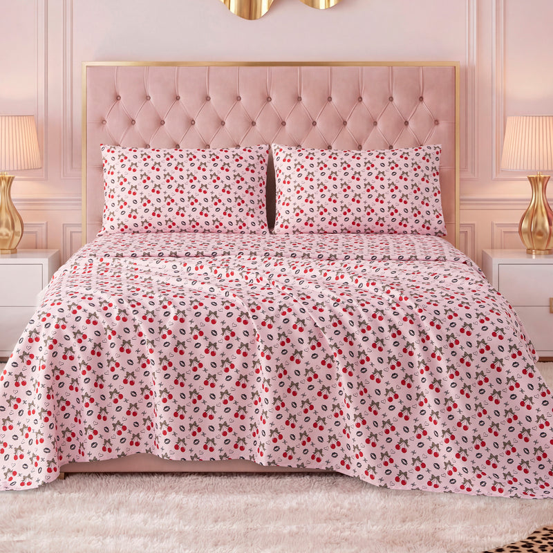 A bed with a tufted pink headboard features a white bedspread with red and black floral designs, paired with a Printed Microfiber Sheet Set, and flanked by white nightstands with gold lamps in a stylish, softly lit bedroom.