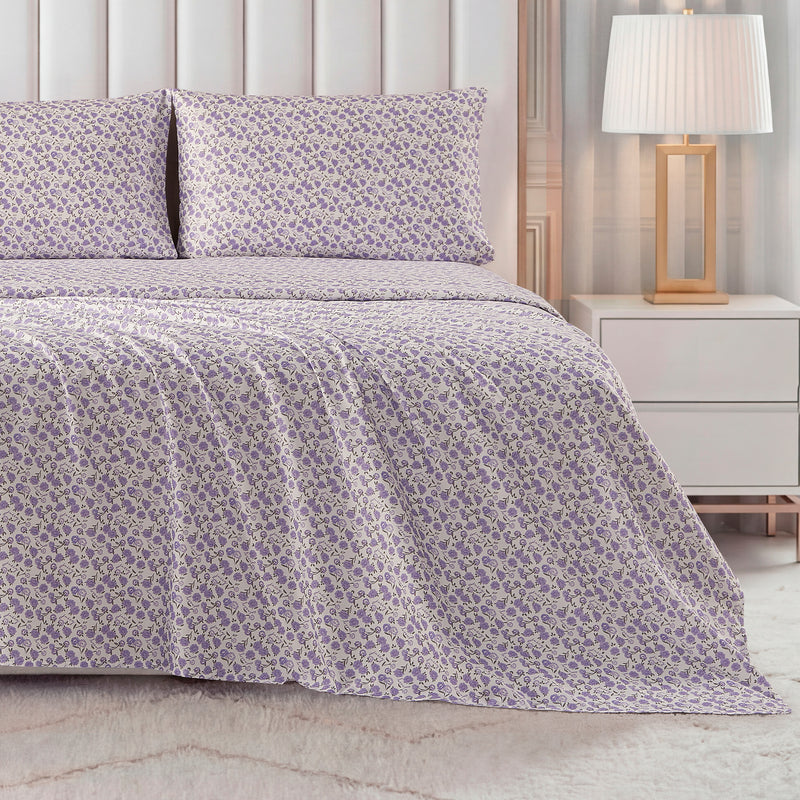 A bed dressed in the Printed Microfiber Sheet Set, featuring a purple and white floral pattern with matching pillowcases, sits beside a white nightstand and gold-base lamp in a warmly lit bedroom.