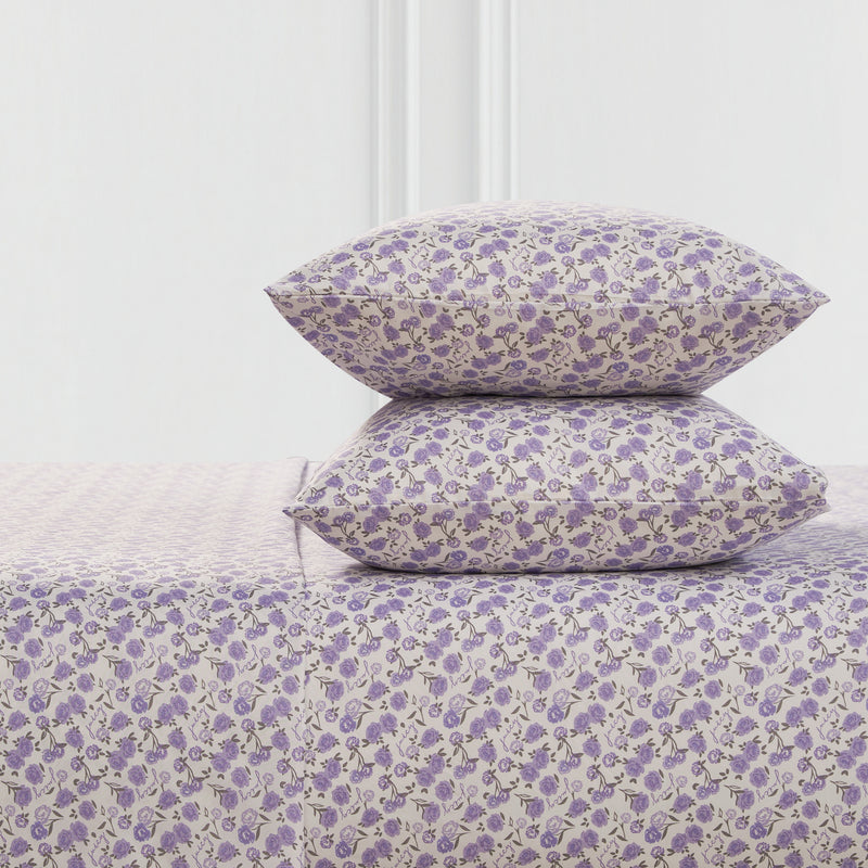 Two pillows and a Printed Microfiber Sheet Set, all with a small purple floral pattern on a light background, are stacked together in front of a minimal white wall.