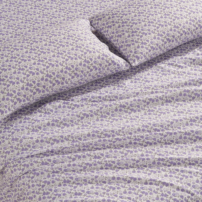A close-up of a bed featuring the Printed Microfiber Sheet Set, showing a pillow and blanket in matching white fabric with a small purple floral pattern.