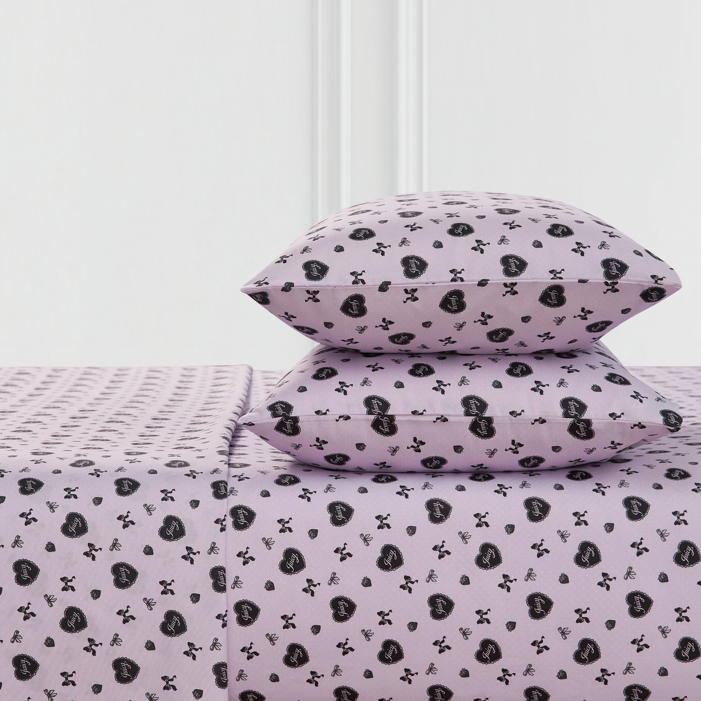Two lavender Printed Microfiber Sheet Set pillows and a matching bedsheet, all with a repeating black heart and crossbones pattern, are neatly arranged against a white wall.