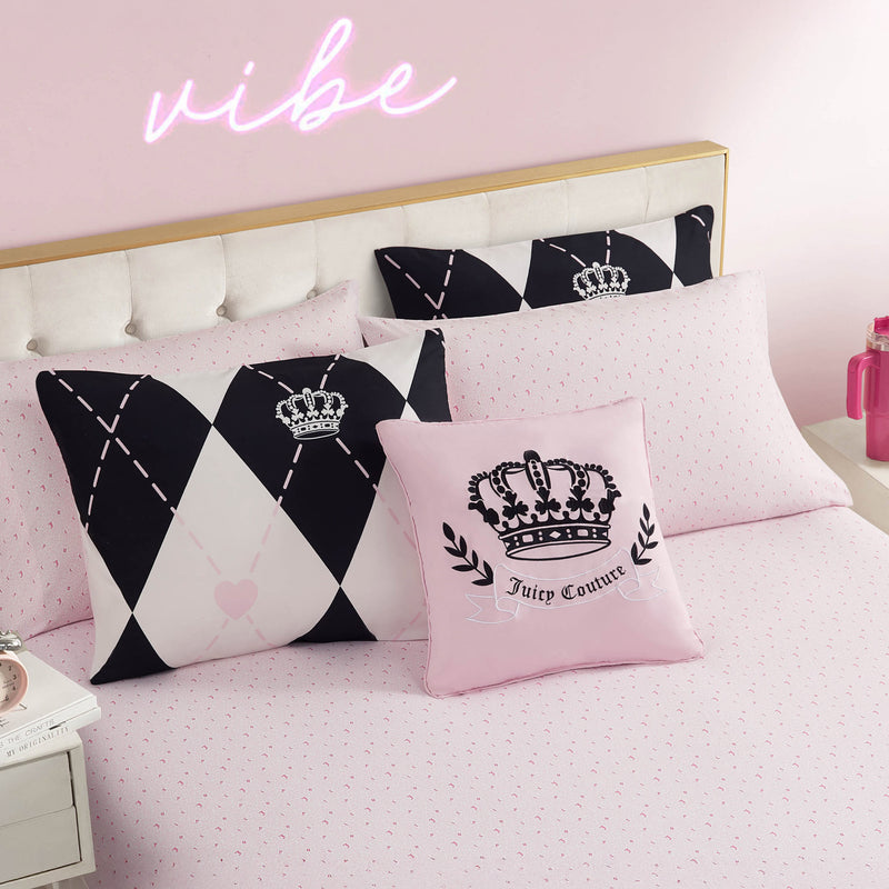 A neatly made bed features the Royal Argyle Reversible Bed in a Bag Bedding Set with pink sheets, black and white diamond-patterned pillows, a Juicy Couture crown pillow, and a glowing neon vibe sign on the pink wall above the headboard.