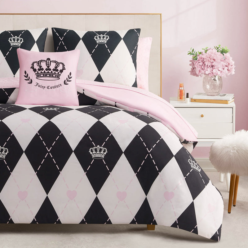 A neatly made bed with the Royal Argyle Reversible Bed in a Bag Bedding Set, featuring a black, white, and pink argyle pattern with crowns and matching Juicy Couture pillow, beside a nightstand with pink flowers in a chic pink-walled room.