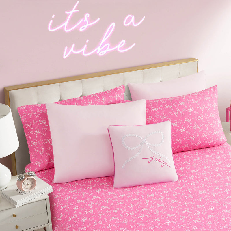 A stylish pink bedroom with the Pearly Bows Reversible Bed in a Bag set featuring bow-patterned sheets, a reversible comforter, a decorative "July" pillow, a white nightstand, and a neon "it's a vibe" wall sign.