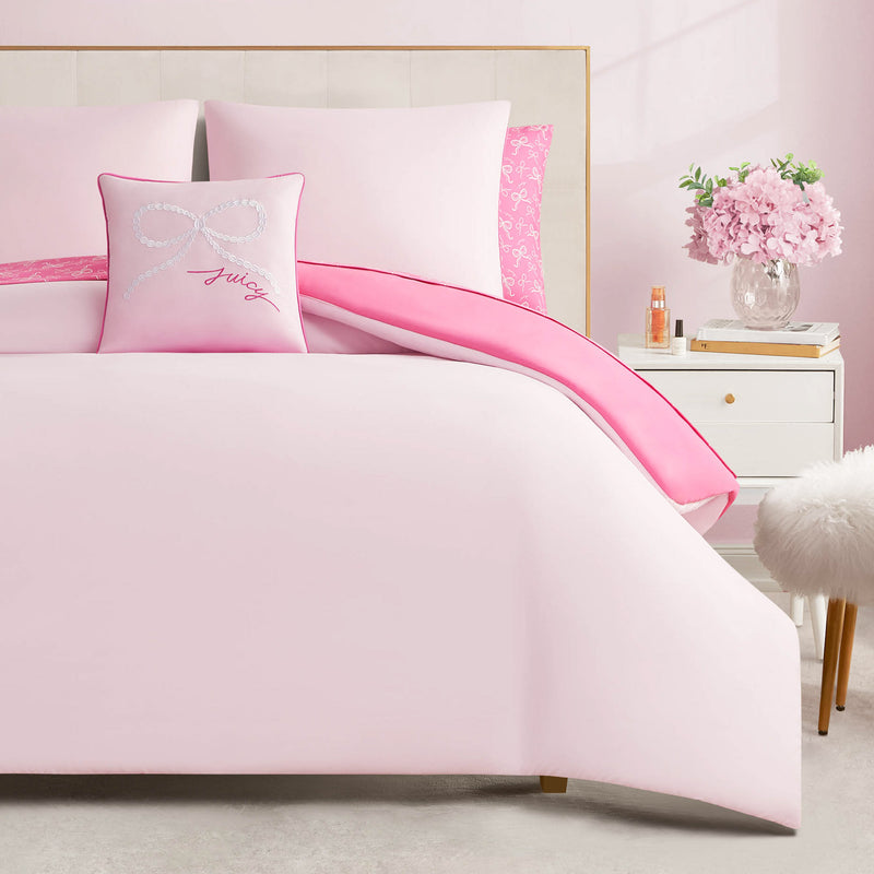 The Pearly Bows Reversible Bed in a Bag Bedding Set creates a soft, feminine look with its light pink bedding and bow pillow. A white nightstand nearby holds pink flowers and perfume, adding designer-inspired charm to the bedroom.