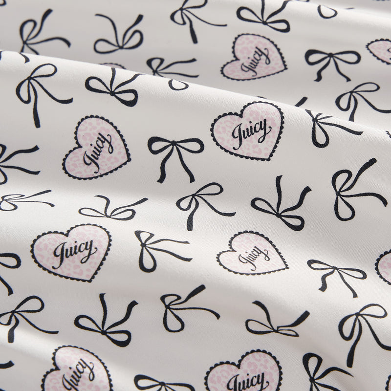 The Printed Microfiber Sheet Set features a white base with black bows and pink polka-dotted hearts outlined in black, each heart labeled "Juicy" in script. The luxury bedding is softly draped with gentle folds.