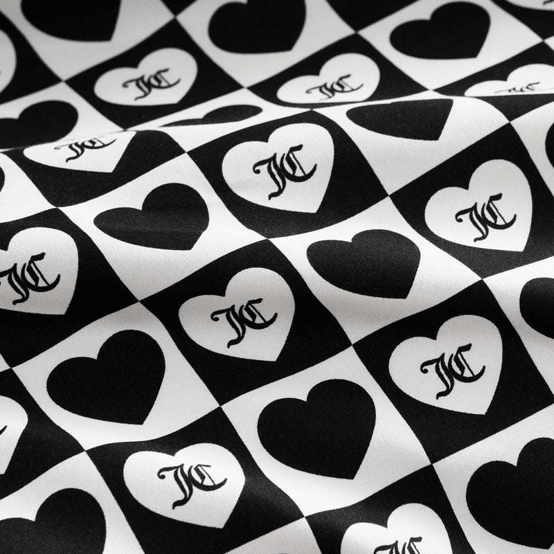 The Printed Microfiber Sheet Set features black and white checkerboard squares with alternating hearts, each displaying a stylized overlapping MC monogram. The luxury bedding surface has a subtle wavy texture.
