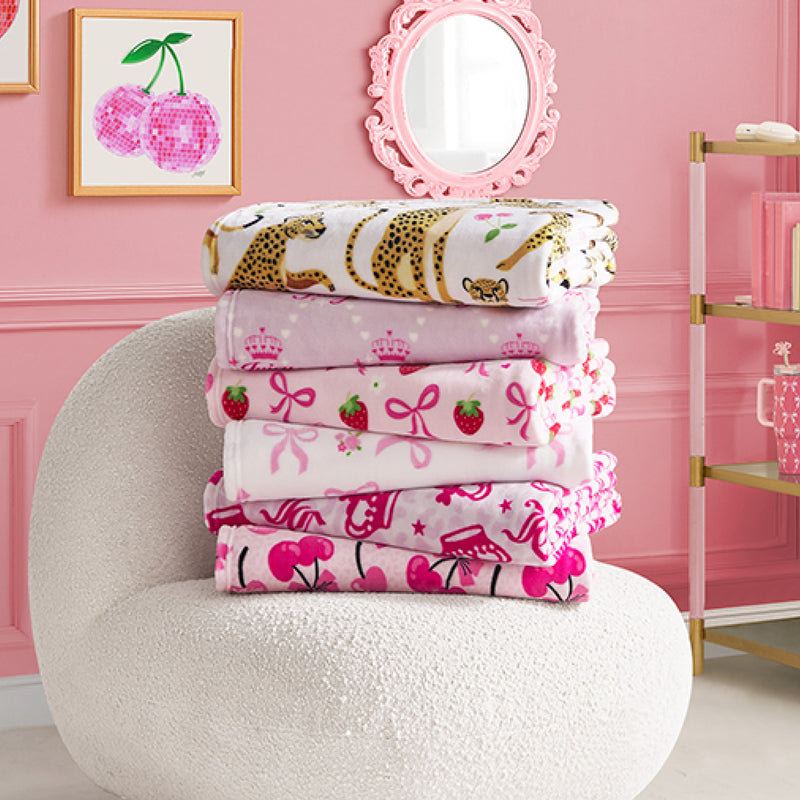 A stack of colorful Kids Plush Throw Blankets, including an ultra-soft Juicy Couture Kids design, is neatly folded on a white textured chair in a pink room. Patterns feature leopards, crowns, cherries, and more; cherry art and a mirror decorate the wall.