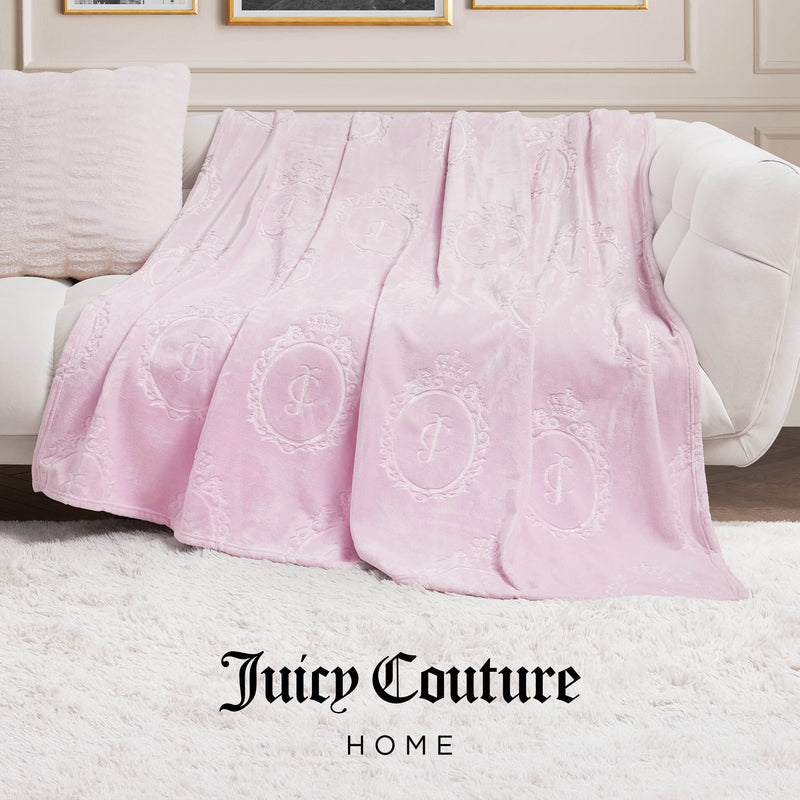 A light pink Plush Oversized Throw Blanket (60" x 90") with embossed logo patterns is draped over a white couch in a chic living room, featuring "Juicy Couture HOME" text at the bottom.