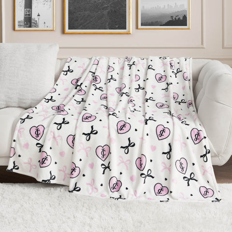 A Plush Oversized Throw Blanket featuring pink hearts, bows, and heart-shaped locks by Juicy Couture is draped over a light sofa in a chic living room with black-and-white photos, adding comfort and style to the space.