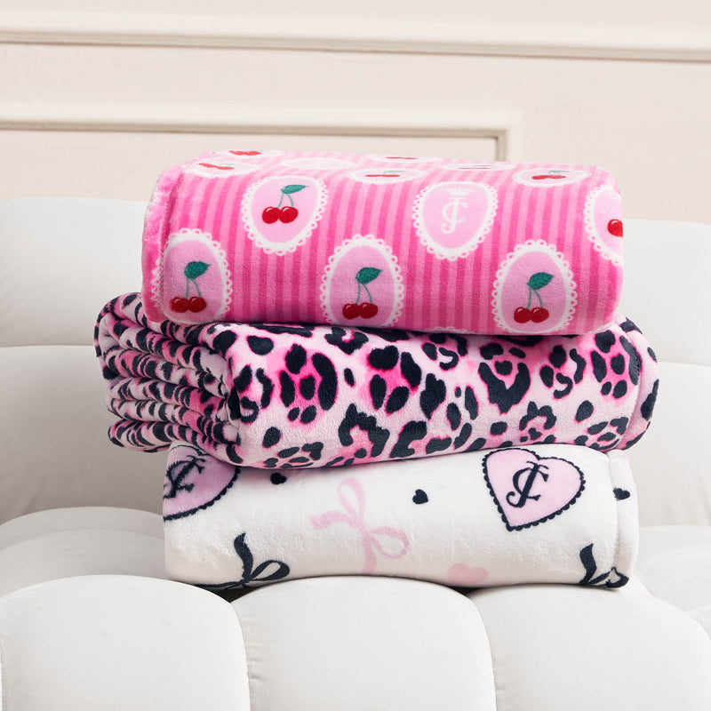Three folded Plush Oversized Throw Blankets stacked on a white cushioned surface: the top is a pink Juicy Couture design with cherries and monograms, the middle has a pink and black leopard print, and the bottom is white with pink hearts and monograms.