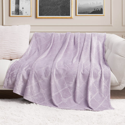 A Plush Oversized Throw Blanket, 60" x 90", in soft lavender with a subtle pattern is draped over a white sofa in a chic living room featuring framed art and a fluffy white rug.