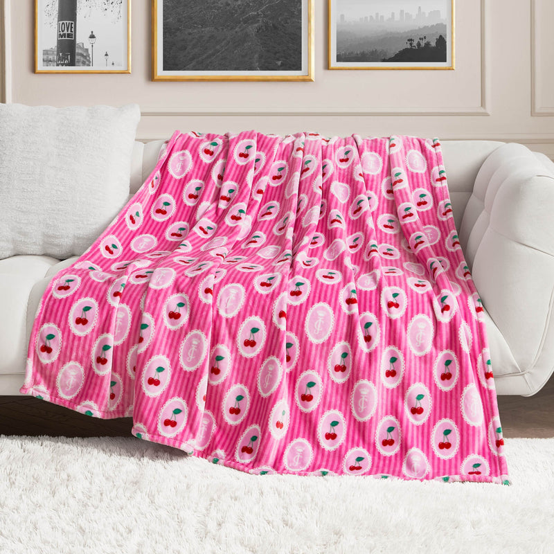 A Plush Oversized Throw Blanket in pink with smiling cherry design is draped over a white couch in a modern living room featuring framed photos and a fuzzy white rug.