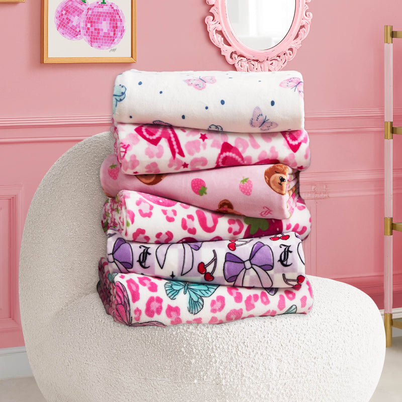 A stack of six Kids Plush Throw Blankets with butterfly, bow, bear, and heart designs sits on a white textured chair in a pink room decorated with wall art and a mirror.