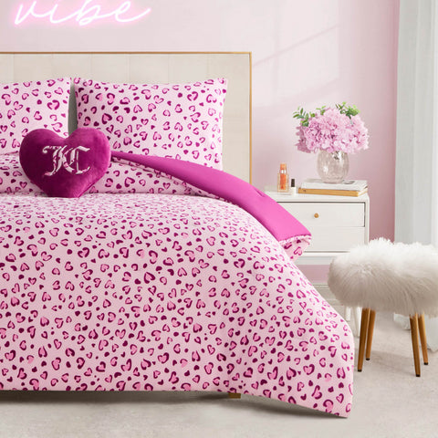 A neatly made bed features the Plush Reversible Leopard Hearts Comforter Set in pink and white, two matching pillows, a decorative heart-shaped purple pillow with "K", a white nightstand with flowers and books, and a fluffy stool in a pink-themed room.