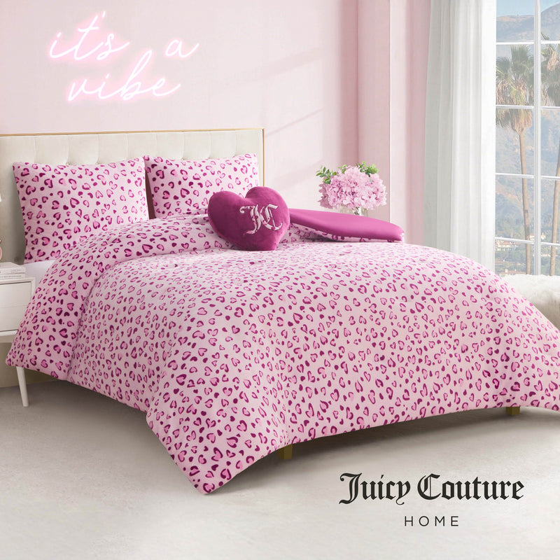 A bedroom featuring the Plush Reversible Leopard Hearts Comforter Set with heart-shaped pillows and matching shams, a neon “it’s a vibe” sign on the pink wall, palm trees outside the window, and the Juicy Couture Home logo in the corner.