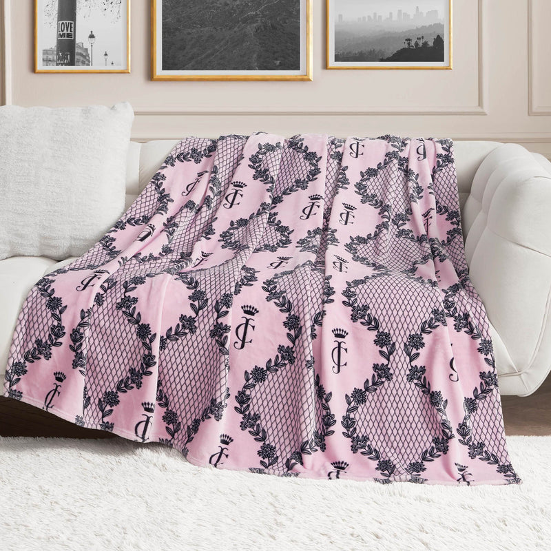A Plush Oversized Throw Blanket by Juicy Couture, pink with a black crown and JC pattern, is draped over a white sofa. Ultra-soft and stylish, it adds flair to the room as framed photos hang on the wall behind.