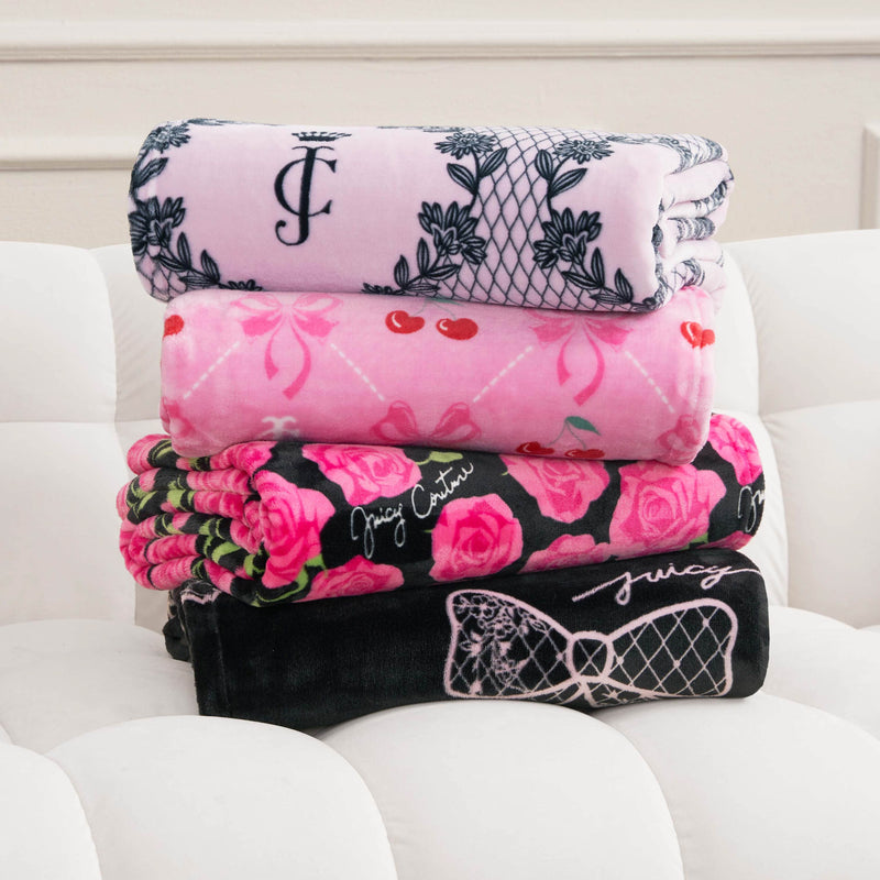 A stack of four folded Plush Oversized Throw Blankets, including a Juicy Couture design in pink and black with floral patterns, cherries, and script accents, is displayed on a white cushioned surface.