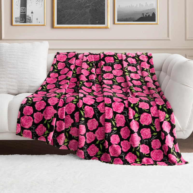 The Plush Oversized Throw Blanket, featuring vibrant pink roses and green leaves on a black background, is draped over a white couch in a bright, elegant living room decorated with framed artwork.