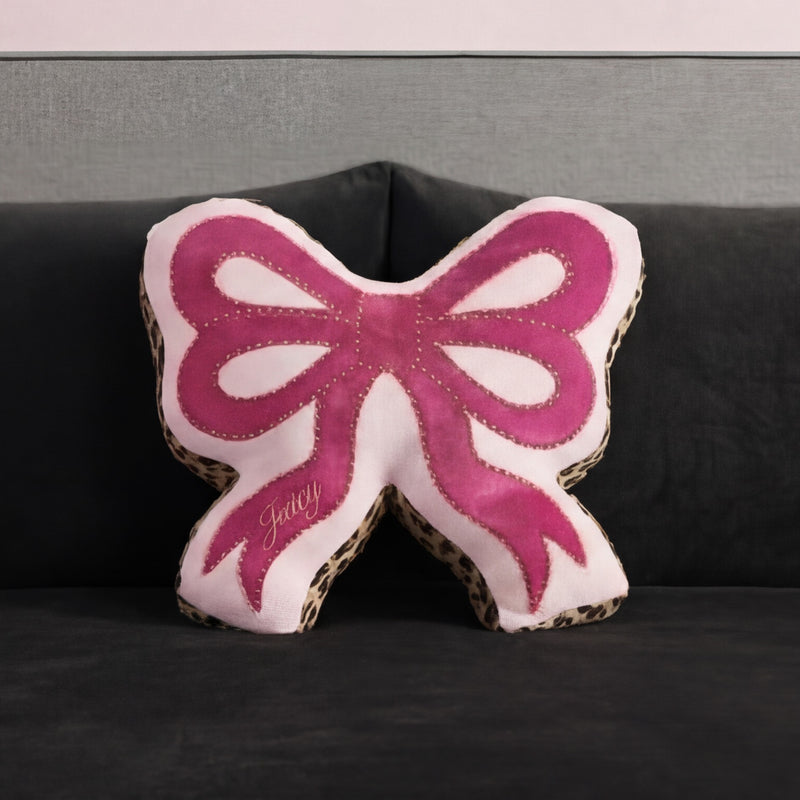 A Reversible Bow Decorative Pillow by Juicy Couture, pink with leopard print edges and "Juicy" embroidery, sits on a dark gray sofa against a matching gray wall.