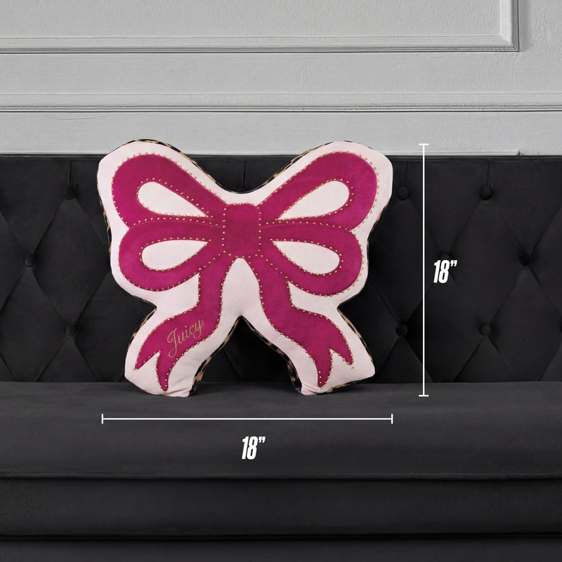 A Reversible Bow Decorative Pillow by Juicy Couture, pink and white with "Juicy" on it, sits on a black tufted sofa. Shaped like a bow, this decorative pillow measures 18" x 18".