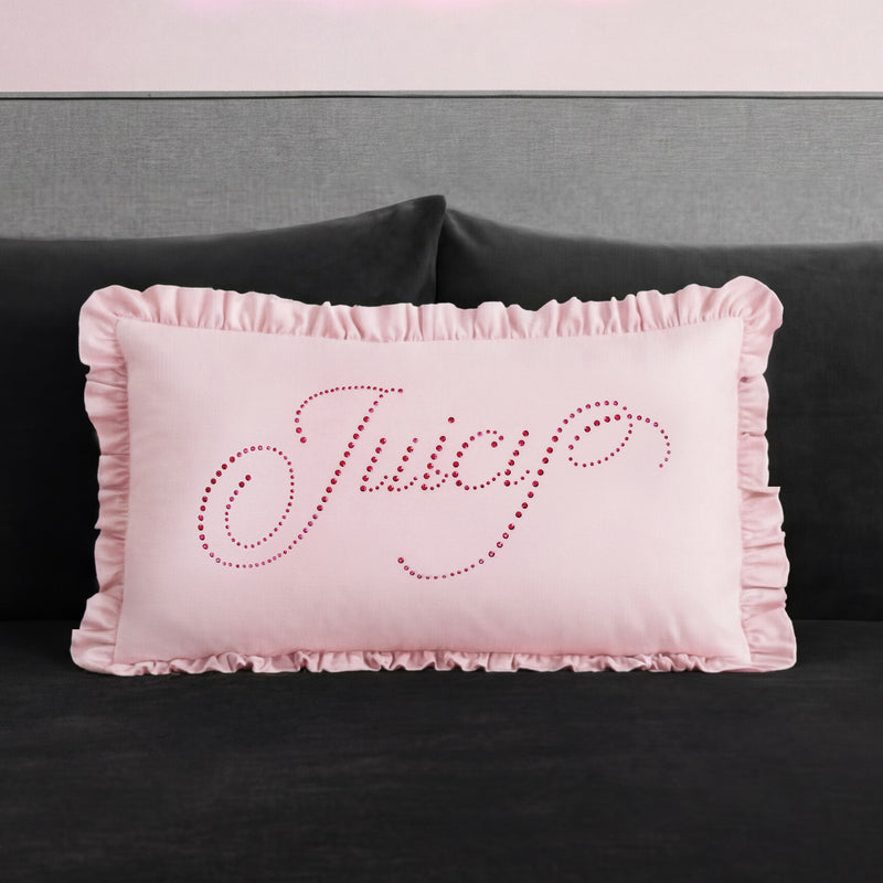 A rectangular pink Ruffle Decorative Pillow with ruffled edges sits on a dark couch. "Juicy" is written in cursive on the pillow with small, shiny pink studs for a glamorous touch.