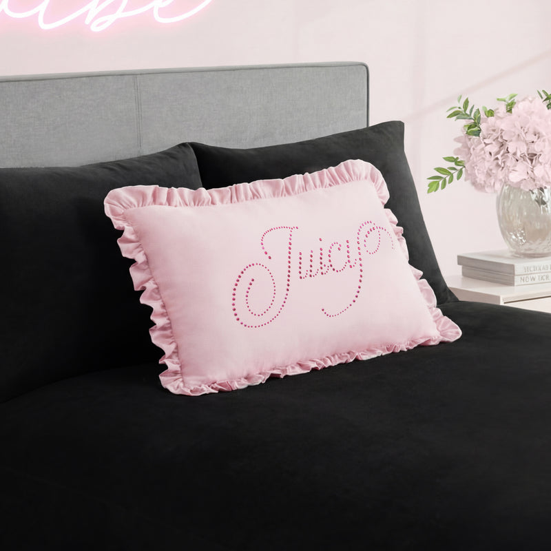 A pink Ruffle Decorative Pillow with rhinestone accents sits on a black bedspread, paired with a gray headboard and a vase of pink flowers on the bedside table in the background.