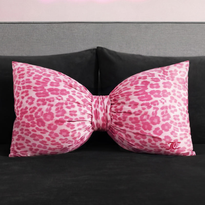 A Bow Decorative Pillow from Juicy Couture, featuring a pink and white leopard print and a small embroidered monogram, rests on a dark gray sofa or bed.
