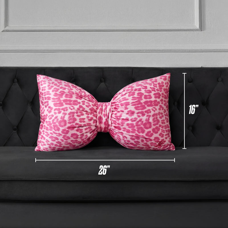 A Bow Decorative Pillow in pink cheetah print, sized 26" x 16", is placed on a black tufted sofa against a gray paneled wall.