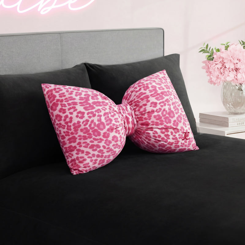 The Bow Decorative Pillow, featuring a large pink cheetah print design, adds a stylish touch to the black bedspread in a chic bedroom with light walls and a vase of pink flowers on the bedside table.