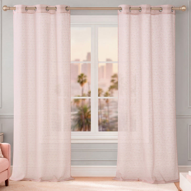Two Aurora Sheer Window Curtains with grommet headers hang from a gold rod in front of a window, showcasing a cityscape and palm trees outside. The room features light walls and a partially visible pink armchair on the left.
