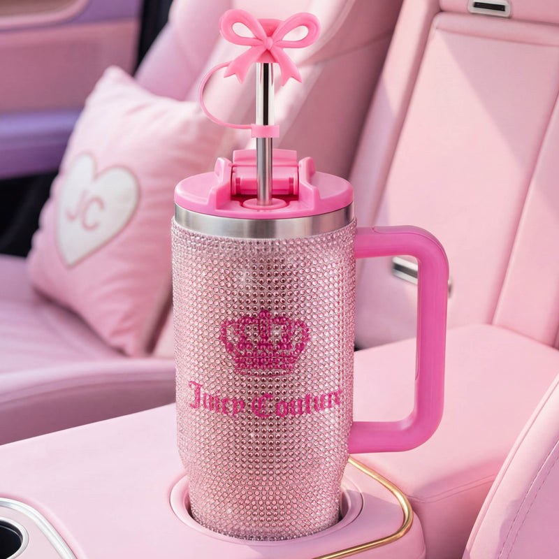 A Diamond Bling Stainless Steel Tumbler with Handle and Straw, 30 oz, adorned with jeweled accents and a bow-shaped straw, sits in the cup holder of a pink car with matching pink seats and a heart-shaped pillow embroidered with JC.