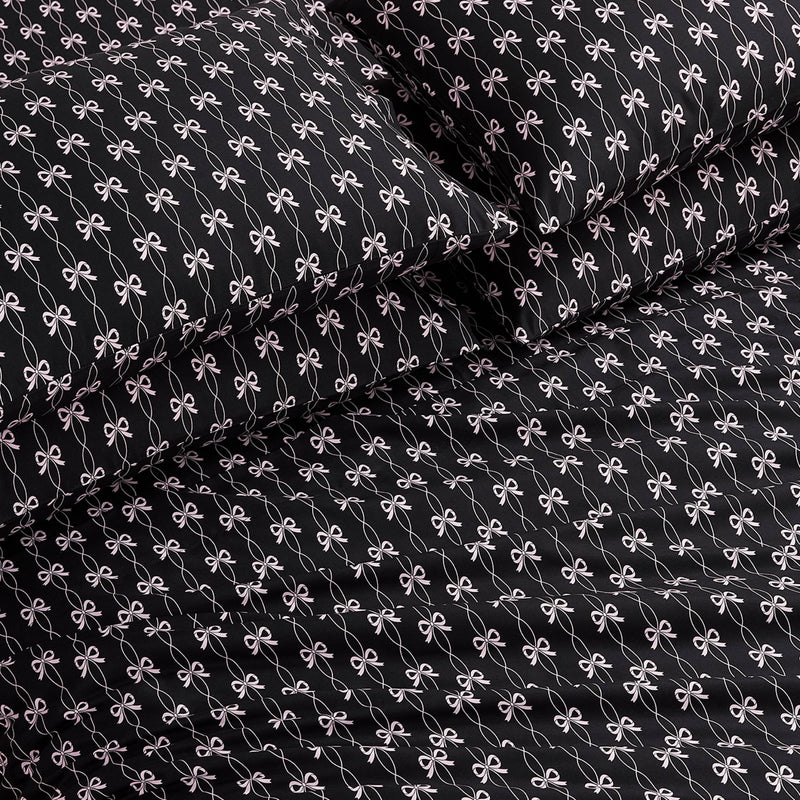 The Microfiber Sheet Set With Extra Pillowcases features black luxury bedding with a repeating small white bow pattern, including a fitted sheet, flat sheet, and two pillowcases, all neatly arranged on a bed.