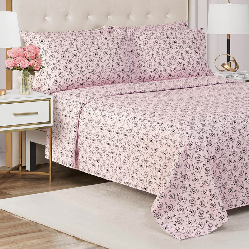 A neatly made bed with a Microfiber Sheet Set With Extra Pillowcases in pink floral patterns and matching pillowcases sits in a modern bedroom featuring a white nightstand, pink flowers, gold accents, and a white table lamp.