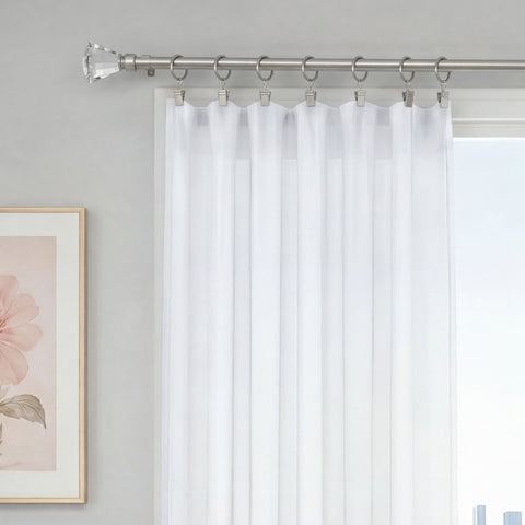 White sheer curtains hang from the Malibu 2-Piece Adjustable Curtain Rod Set, which features a telescoping, adjustable design. The curtains are elegantly suspended by rings and clips to cover a window. On the left wall, a framed flower artwork is visible, contributing to the room’s bright and minimalistic decor.