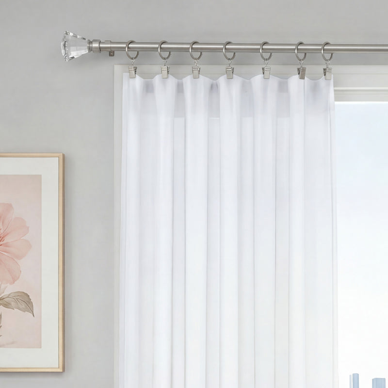 White sheer curtains hang from the Malibu 2-Piece Adjustable Curtain Rod Set, which features a telescoping, adjustable design. The curtains are elegantly suspended by rings and clips to cover a window. On the left wall, a framed flower artwork is visible, contributing to the room’s bright and minimalistic decor.