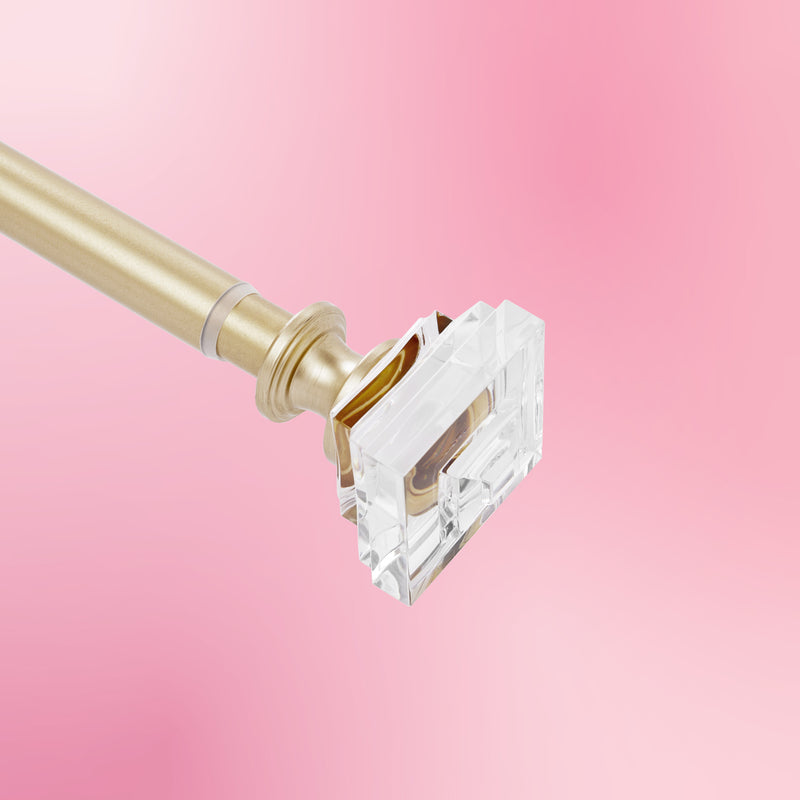 The Rodeo Adjustable Curtain Rod, featuring a gold finish and clear rectangular acrylic finial, is displayed against a pink gradient background, bringing Juicy Couture-inspired elegance to your window décor.