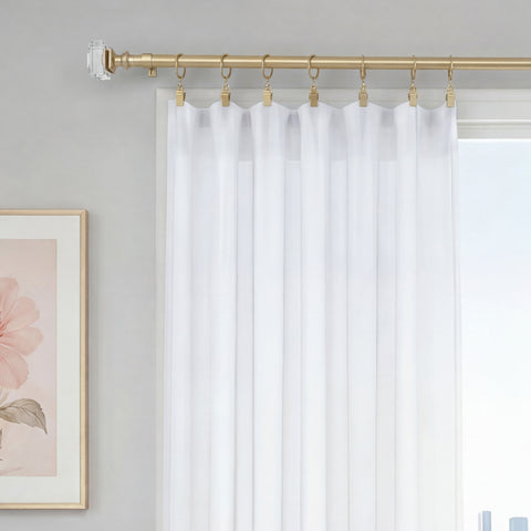 The Rodeo Adjustable Curtain Rod in a gold finish with clear finials supports sheer white curtains, adding an elegant touch to your window decor. A glimpse of the window and a framed pink floral artwork on a neutral wall are also visible.