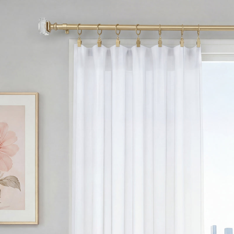 The Rodeo Adjustable Curtain Rod in a gold finish with clear finials supports sheer white curtains, adding an elegant touch to your window decor. A glimpse of the window and a framed pink floral artwork on a neutral wall are also visible.