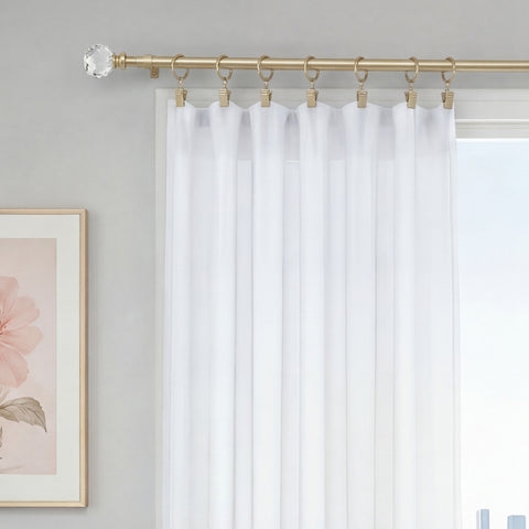 A white sheer curtain is displayed on the Astoria 2-Piece Adjustable Curtain Rod Set, which features gold finish with decorative acrylic accents and rings, beside a window. A portion of a framed floral artwork hangs on the light gray wall nearby.