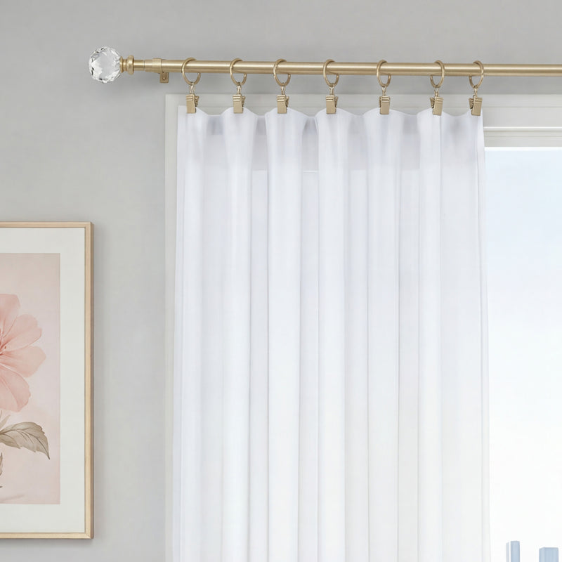 A white sheer curtain is displayed on the Astoria 2-Piece Adjustable Curtain Rod Set, which features gold finish with decorative acrylic accents and rings, beside a window. A portion of a framed floral artwork hangs on the light gray wall nearby.