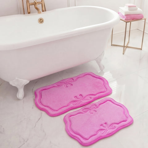 A white clawfoot bathtub stands next to two pink Quilted Memory Foam Bath Mats, both with bow designs, on a white marble floor. A gold stand with folded towels is in the background.