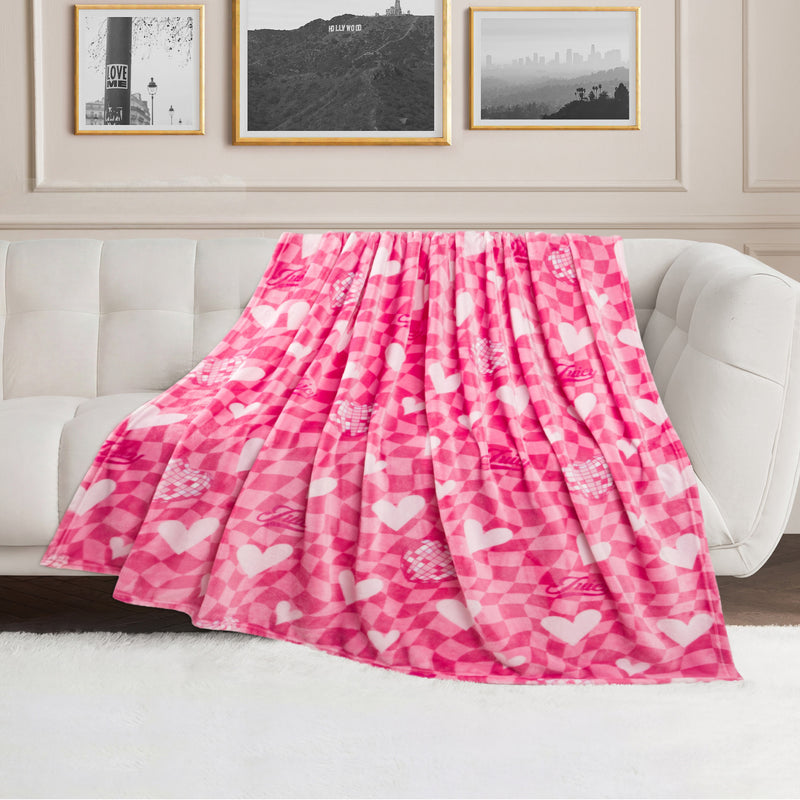 A Plush Oversized Throw Blanket, 60″ x 70″, featuring a pink and white heart pattern, is draped over a white sofa in a modern living room with framed art on the wall.