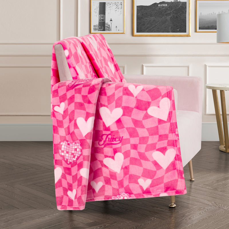 A Plush Oversized Throw Blanket, 60″ x 70″, with a pink heart pattern is draped over a pale pink armchair in a stylish, modern living room featuring framed wall art and wood flooring.