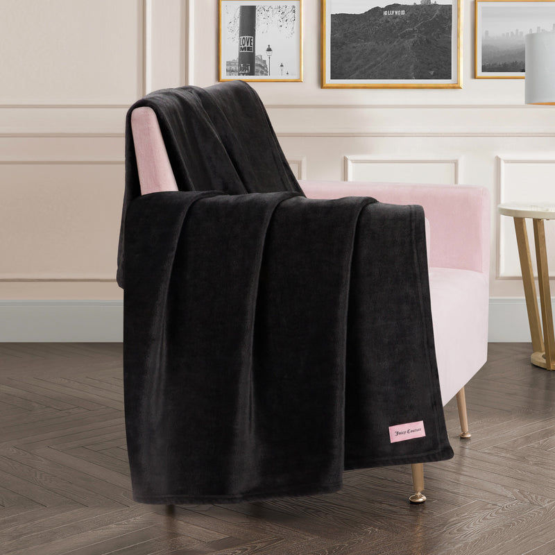 A Plush Oversized Throw Blanket, 60″ x 70″, ultra-soft and cozy, is draped over a light pink armchair beside a gold side table, with framed wall art in the background.