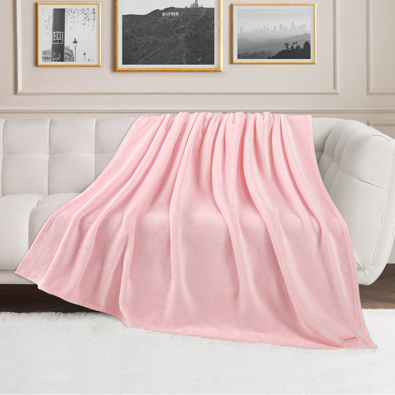A Plush Oversized Throw Blanket, 60″ x 70″, in a soft light pink hue is draped over a white sofa in a stylish living room adorned with framed pictures and a fluffy white rug.