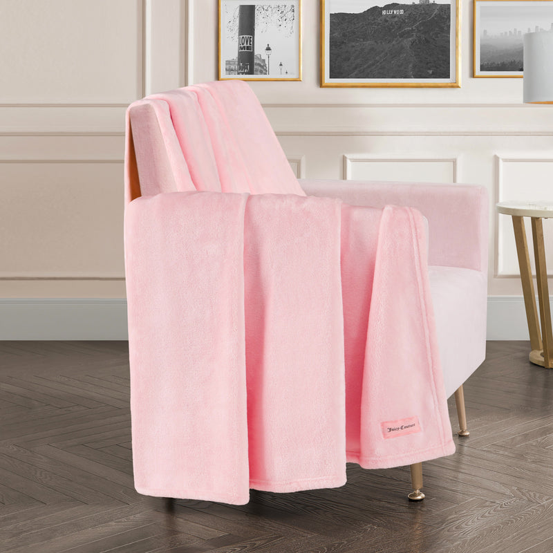 A Plush Oversized Throw Blanket, 60″ x 70″, in soft pink is draped over a matching pink armchair in a stylish room with white paneled walls, framed art, a small round side table, and dark wood herringbone flooring.