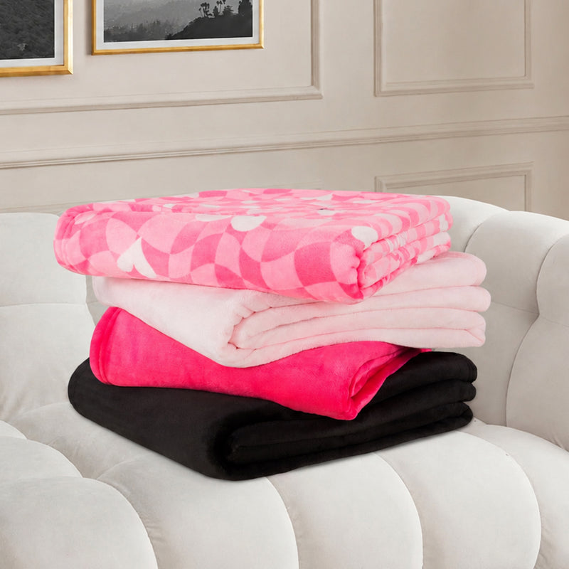 Four folded blankets rest on a white tufted sofa: one pink with heart designs, one light pink, a Plush Oversized Throw Blanket (60″ x 70″) in bright pink, and one black. Two framed pictures hang on the beige wall in the background.
