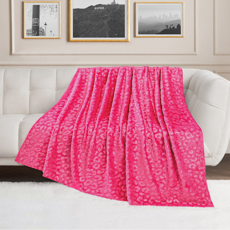 An Embossed Plush Oversized Throw Blanket in bright pink leopard print is draped over a white sofa in a modern living room with framed photos and a white rug.