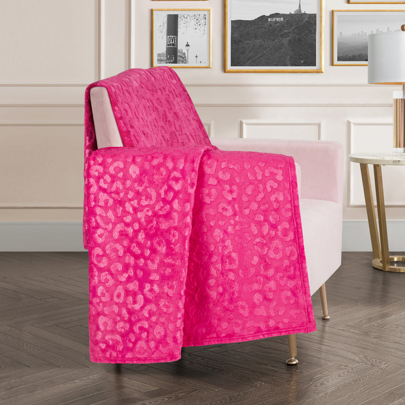 An Embossed Plush Oversized Throw Blanket in vibrant pink with a leopard-print design is draped over a light pink armchair in a stylish living room featuring framed wall art and a gold-legged side table.